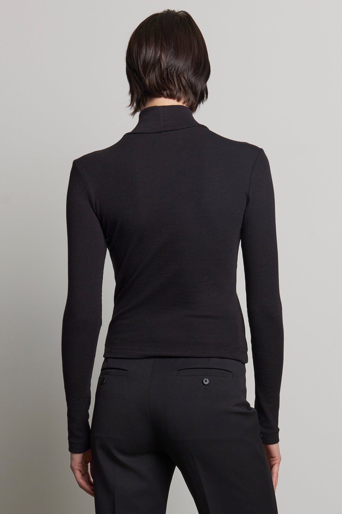 A person with short dark hair is wearing the HELMS TEE, a long-sleeved black mock neck top by Velvet by Jenny Graham, along with black ribbed textured pants, seen from the back against a light gray background.-38318596948161