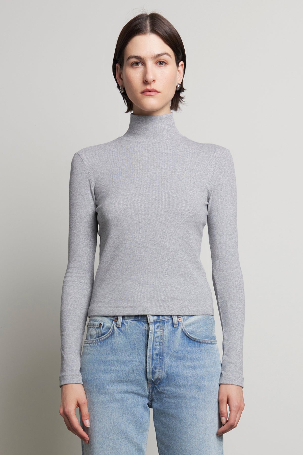 A person wearing the HELMS TEE by Velvet by Jenny Graham and blue jeans stands against a plain background, with the ribbed texture of their mock neck gray turtleneck adding subtle depth to the minimalist scene.-38318596718785