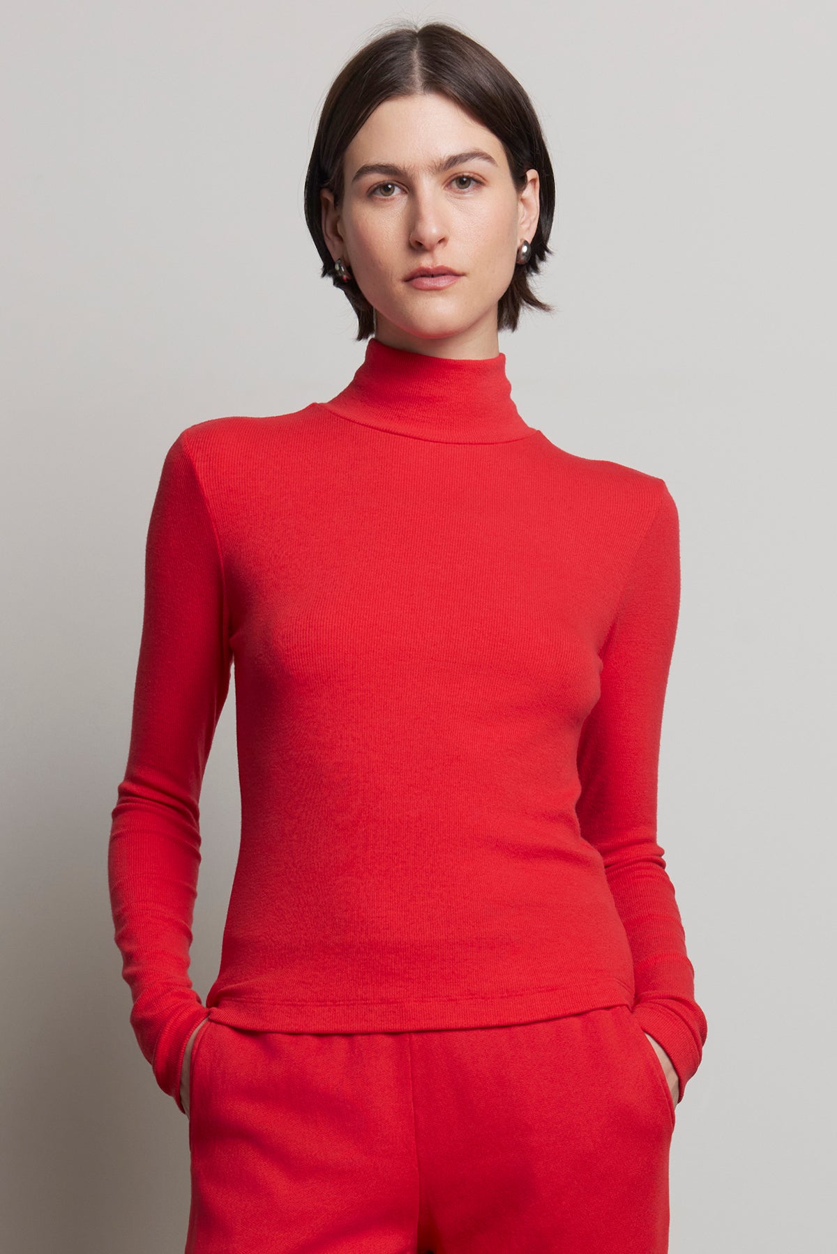   Person wearing the Velvet by Jenny Graham HELMS TEE in red, featuring a mock neck and ribbed texture, paired with matching red pants, stands against a neutral background. 