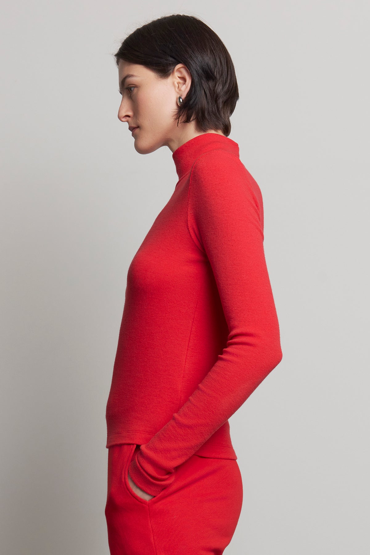   A person in profile is wearing the HELMS TEE, a red turtleneck with a subtle ribbed texture from Velvet by Jenny Graham, paired with pants and standing against a plain background. 