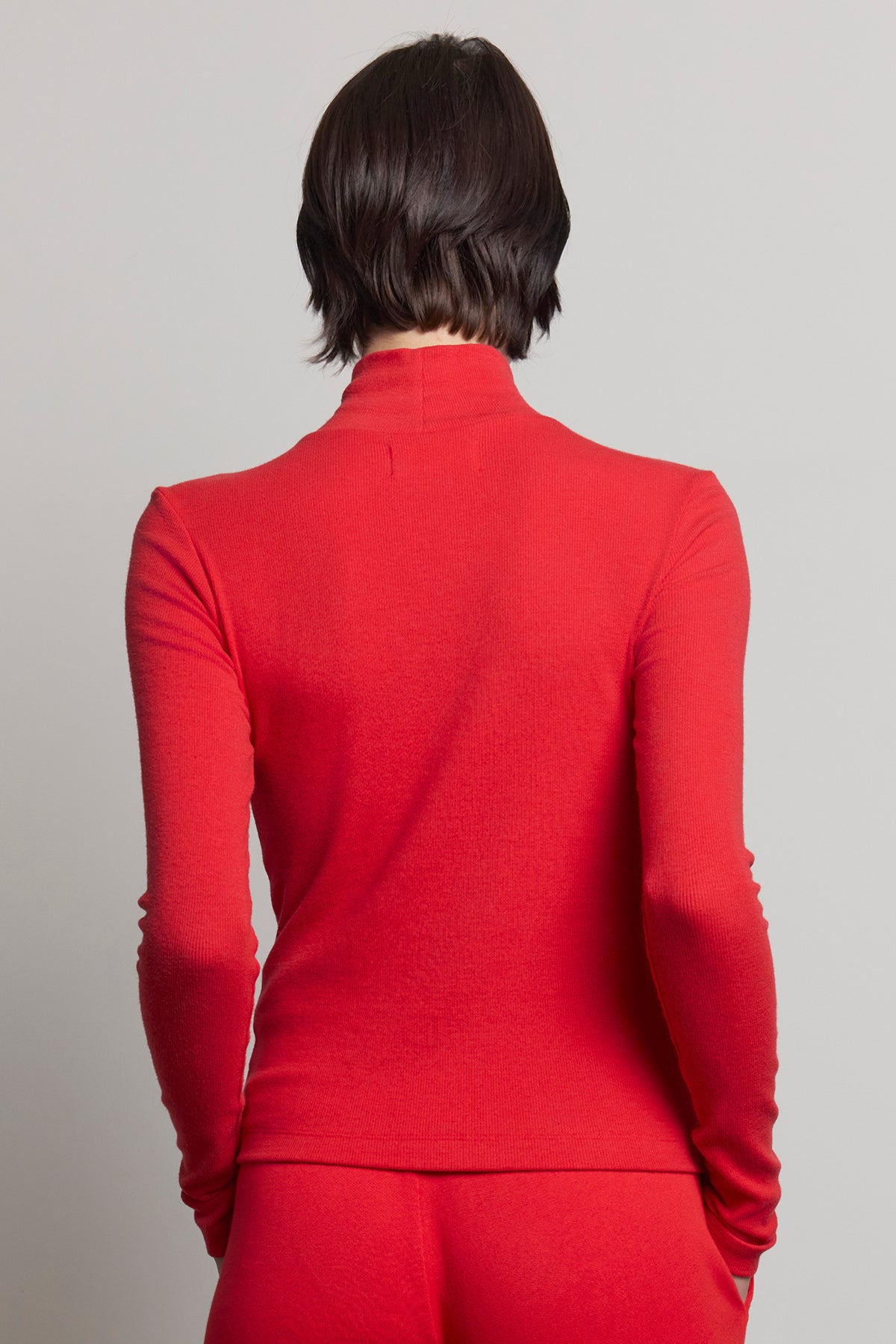   A person with short brown hair is wearing the HELMS TEE, a long-sleeved, red turtleneck sweater from Velvet by Jenny Graham, featuring a ribbed texture. They are facing away from the camera against a plain gray background. 