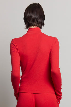 A person with short brown hair is wearing the HELMS TEE, a long-sleeved, red turtleneck sweater from Velvet by Jenny Graham, featuring a ribbed texture. They are facing away from the camera against a plain gray background.