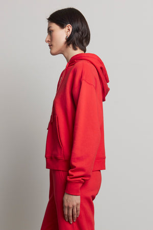 A person wearing an oversized OJAI HOODIE in red from the brand Velvet by Jenny Graham, paired with organic cotton pants, stands in profile against a plain background.