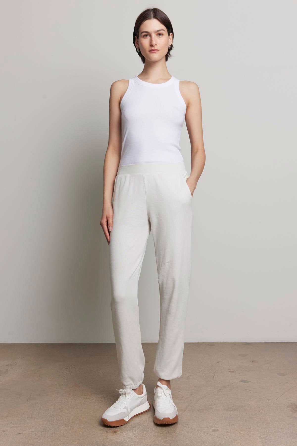 A person is standing against a plain background, dressed in a white tank top and the ZUMA SWEATPANT from Velvet by Jenny Graham, featuring off-white organic cotton material with an elastic waist and pockets, paired with white sneakers, looking at the camera with a neutral expression.-37817332629697