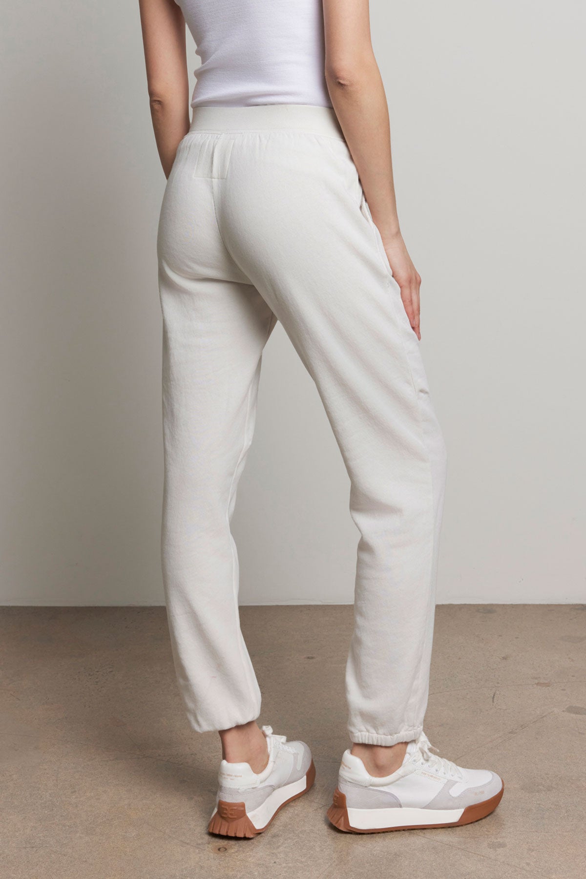 Someone in a white tank top and Velvet by Jenny Graham's ZUMA SWEATPANT stands facing away, highlighting the back of their outfit with its elastic waist. They are wearing white sneakers with tan soles.-37817313165505