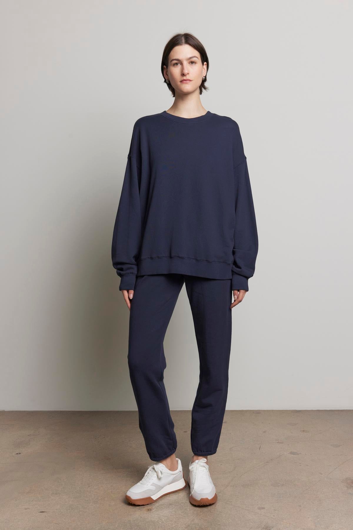 A person stands against a plain background wearing the YNEZ SWEATSHIRT in navy blue from Velvet by Jenny Graham, paired with matching pants and white sneakers.-37817352650945