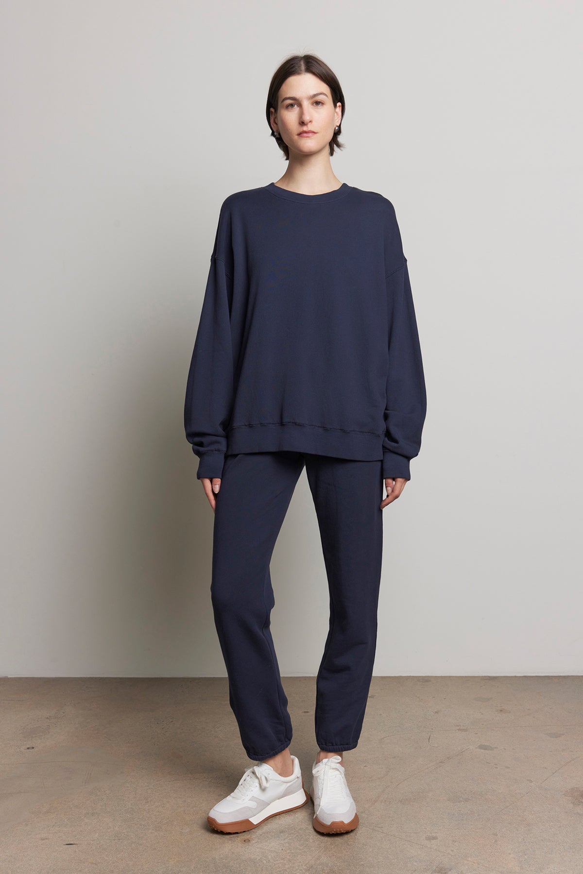   A person wearing a navy sweatshirt and ZUMA SWEATPANT joggers from Velvet by Jenny Graham, made from organic cotton and paired with white sneakers, stands against a plain light gray wall. 