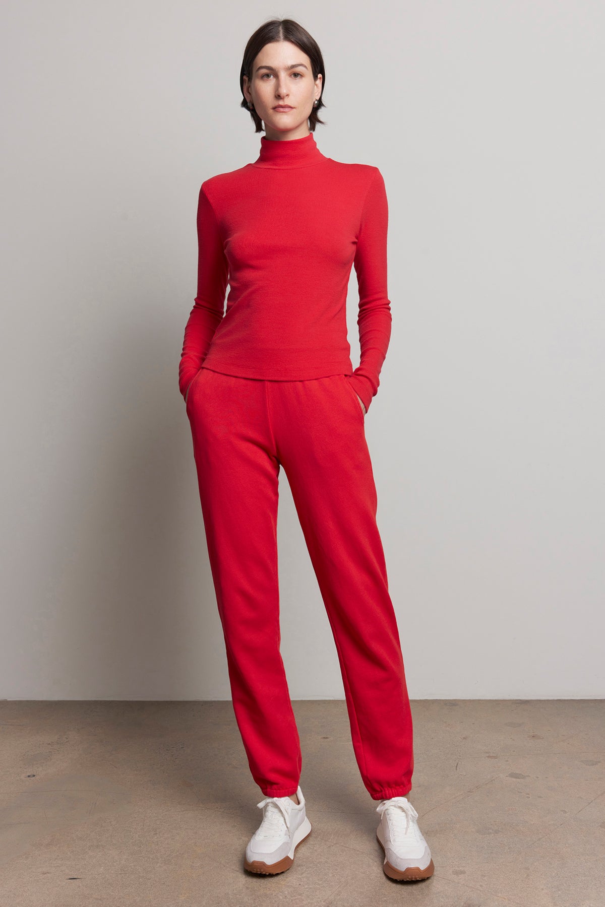   A person in a red turtleneck and matching ZUMA SWEATPANT from Velvet by Jenny Graham stands with hands in pockets, wearing white sneakers. Neutral background highlights this nod to sustainable fashion. 