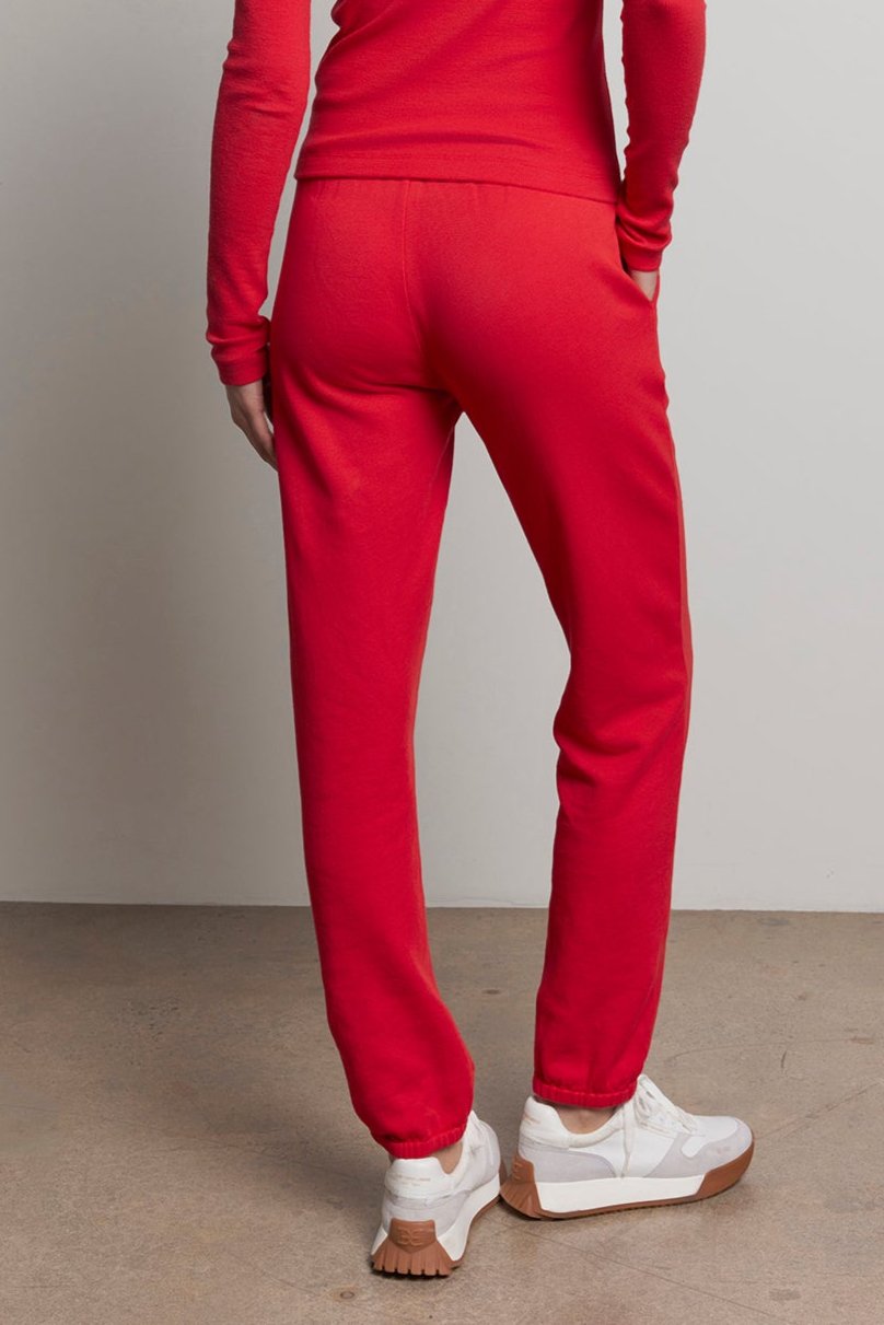   A person wearing a red long-sleeve top and the ZUMA SWEATPANT by Velvet by Jenny Graham stands on a concrete floor, facing away. White sneakers provide contrast to the outfit, embodying sustainable fashion. 