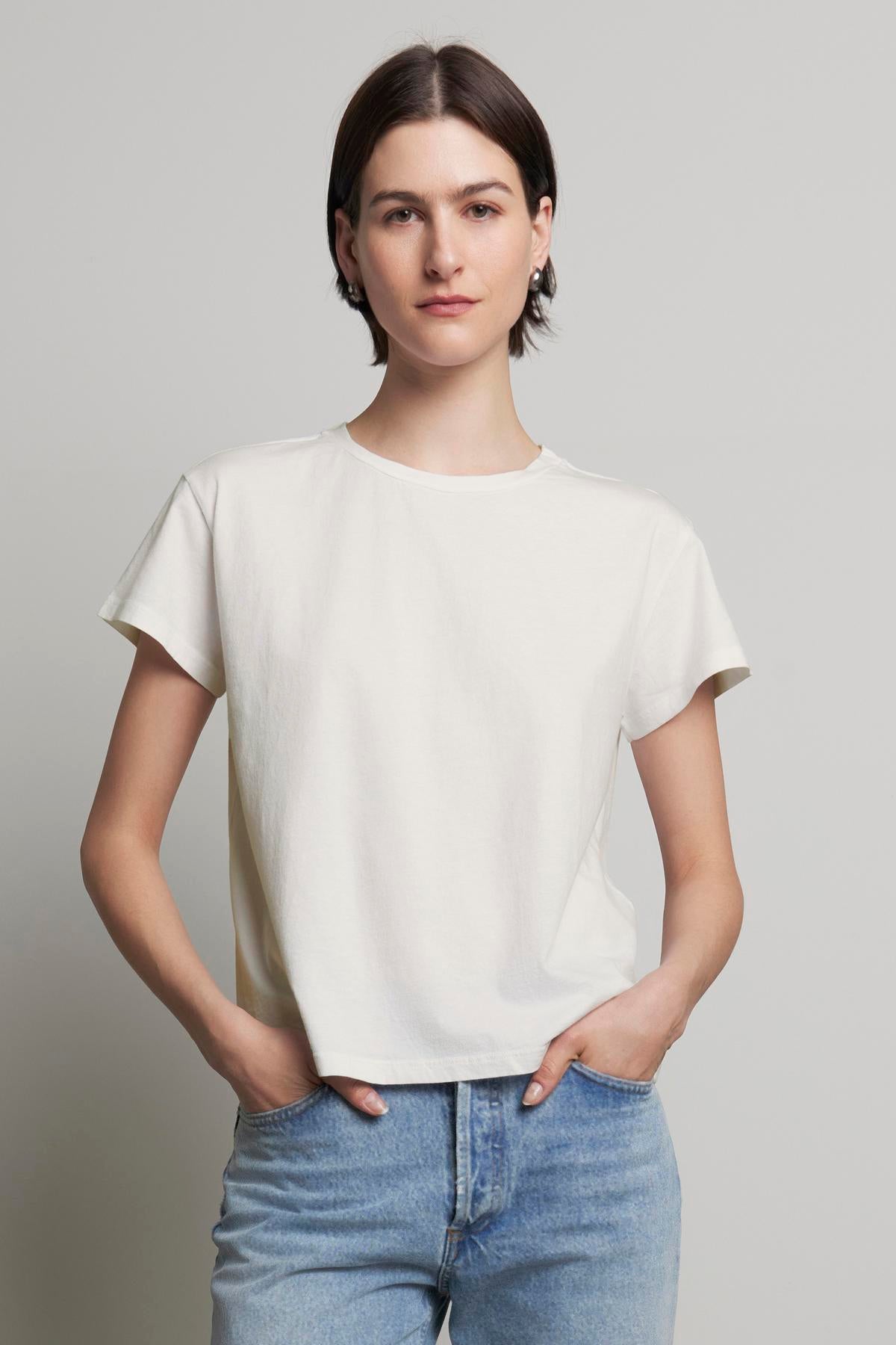   A person with short, dark hair is wearing the TOPANGA TEE by Velvet by Jenny Graham, made of organic cotton, paired with relaxed-fit classic blue jeans, standing with hands in pockets against a plain backdrop. 