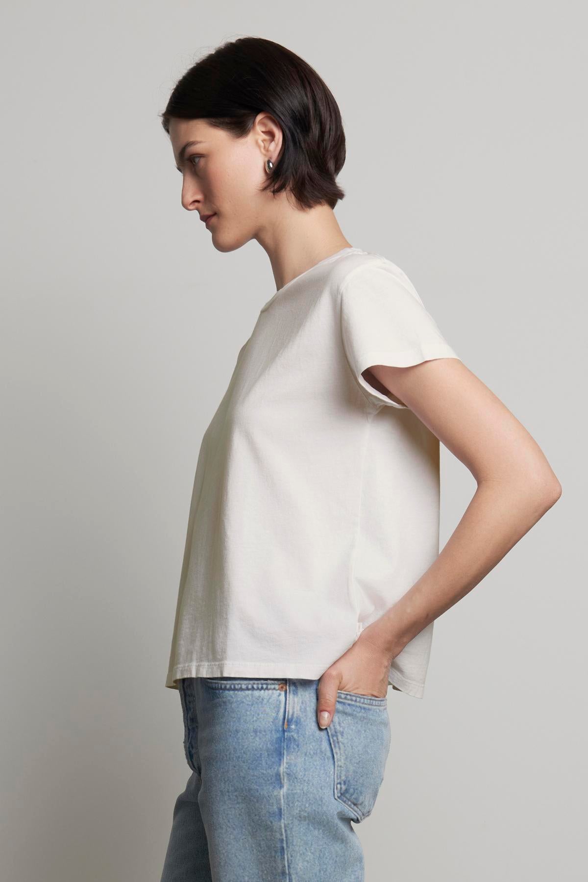   A person with short hair is standing sideways, wearing a relaxed-fit classic TOPANGA TEE from Velvet by Jenny Graham, made from organic cotton, paired with blue jeans and their left hand in their pocket. 