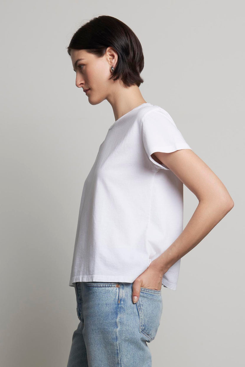 A person with short dark hair stands in profile wearing a relaxed-fit TOPANGA TEE by Velvet by Jenny Graham and blue jeans against a plain background.