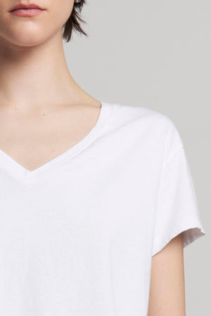 A person in a white VENICE TEE by Velvet by Jenny Graham stands against a neutral background, partially visible from the shoulders up. The organic cotton V-neck T-shirt nods subtly to classic 90s silhouettes.