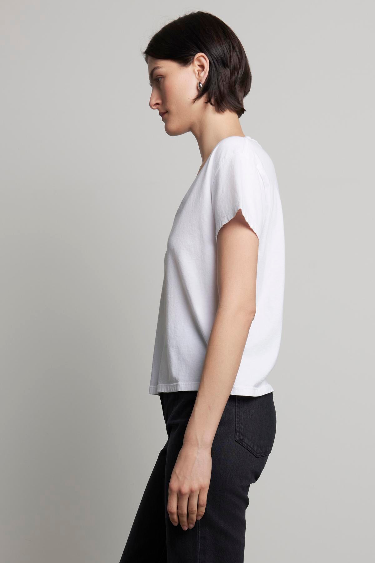   A person with short hair wears the Velvet by Jenny Graham VENICE TEE, a white organic cotton T-shirt with a subtle v-neckline, paired with black pants. They're standing in profile against a plain background, showcasing an outfit inspired by 90s silhouettes. 