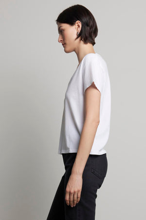 A person with short hair wears the Velvet by Jenny Graham VENICE TEE, a white organic cotton T-shirt with a subtle v-neckline, paired with black pants. They're standing in profile against a plain background, showcasing an outfit inspired by 90s silhouettes.