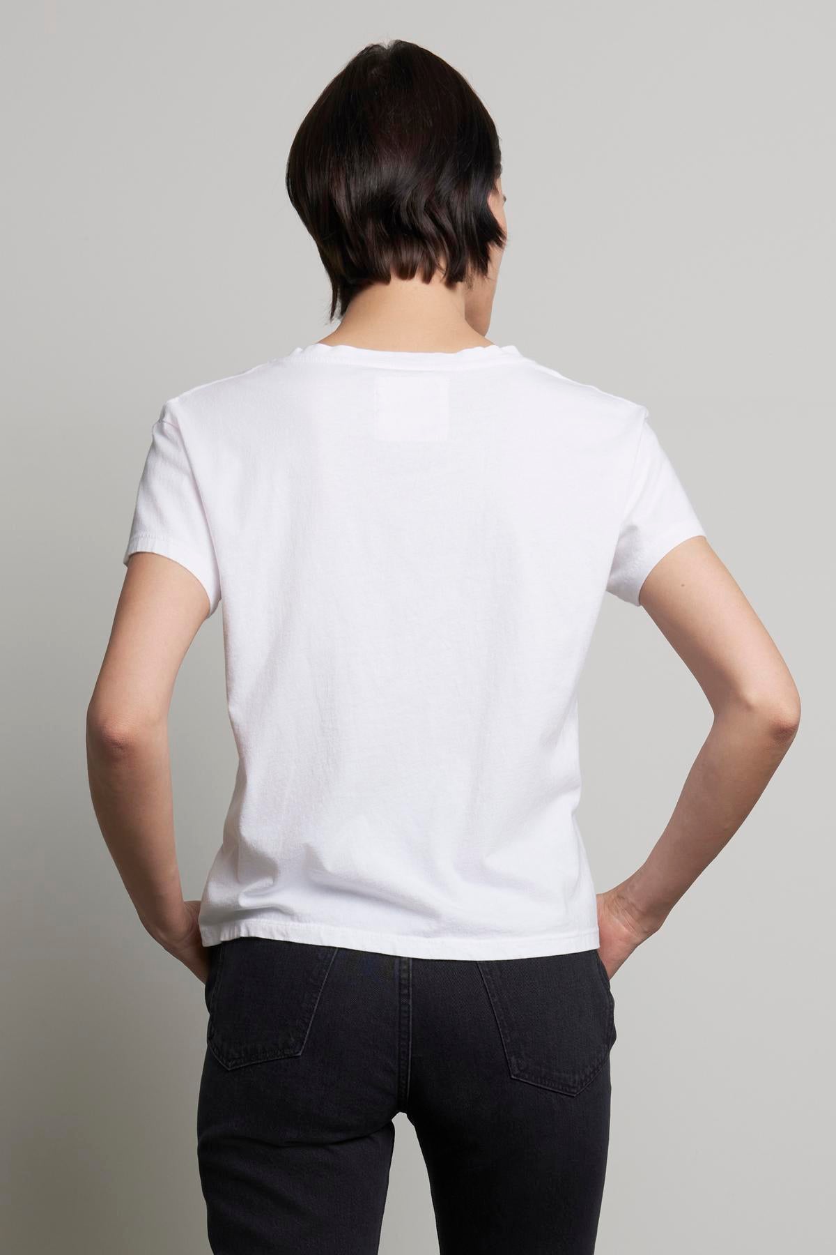   A person with short dark hair, dressed in the VENICE TEE from Velvet by Jenny Graham—a laid-back, chic white organic cotton t-shirt—pairs it with black jeans. They're photographed from behind against a plain background. 