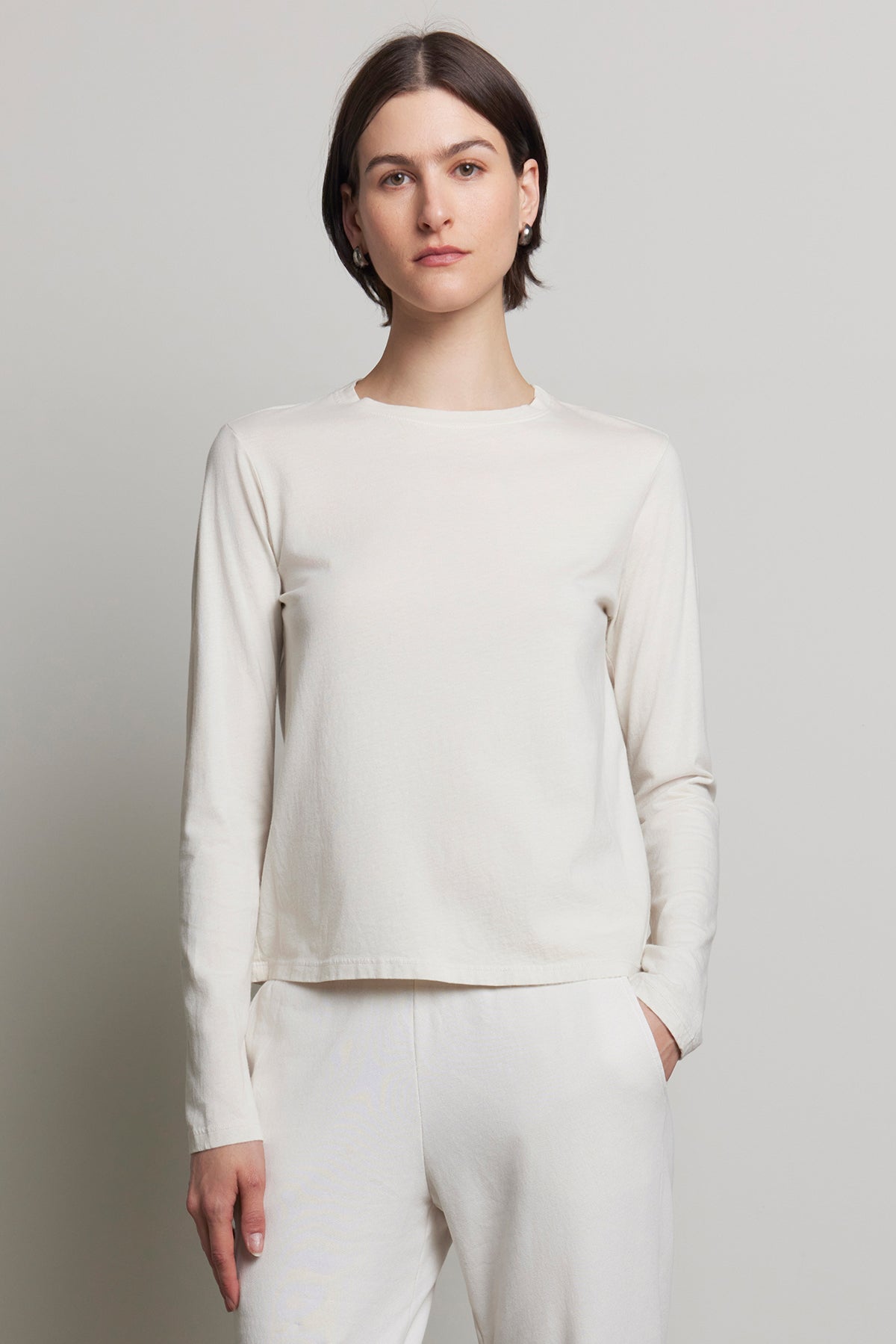   A person stands against a neutral background wearing the VICENTE TEE and white pants, crafted from organic cotton. The plain white long-sleeve shirt by Velvet by Jenny Graham embodies a relaxed fit for an effortlessly chic, seasonless style. 