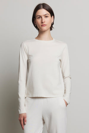 A person stands against a neutral background wearing the VICENTE TEE and white pants, crafted from organic cotton. The plain white long-sleeve shirt by Velvet by Jenny Graham embodies a relaxed fit for an effortlessly chic, seasonless style.