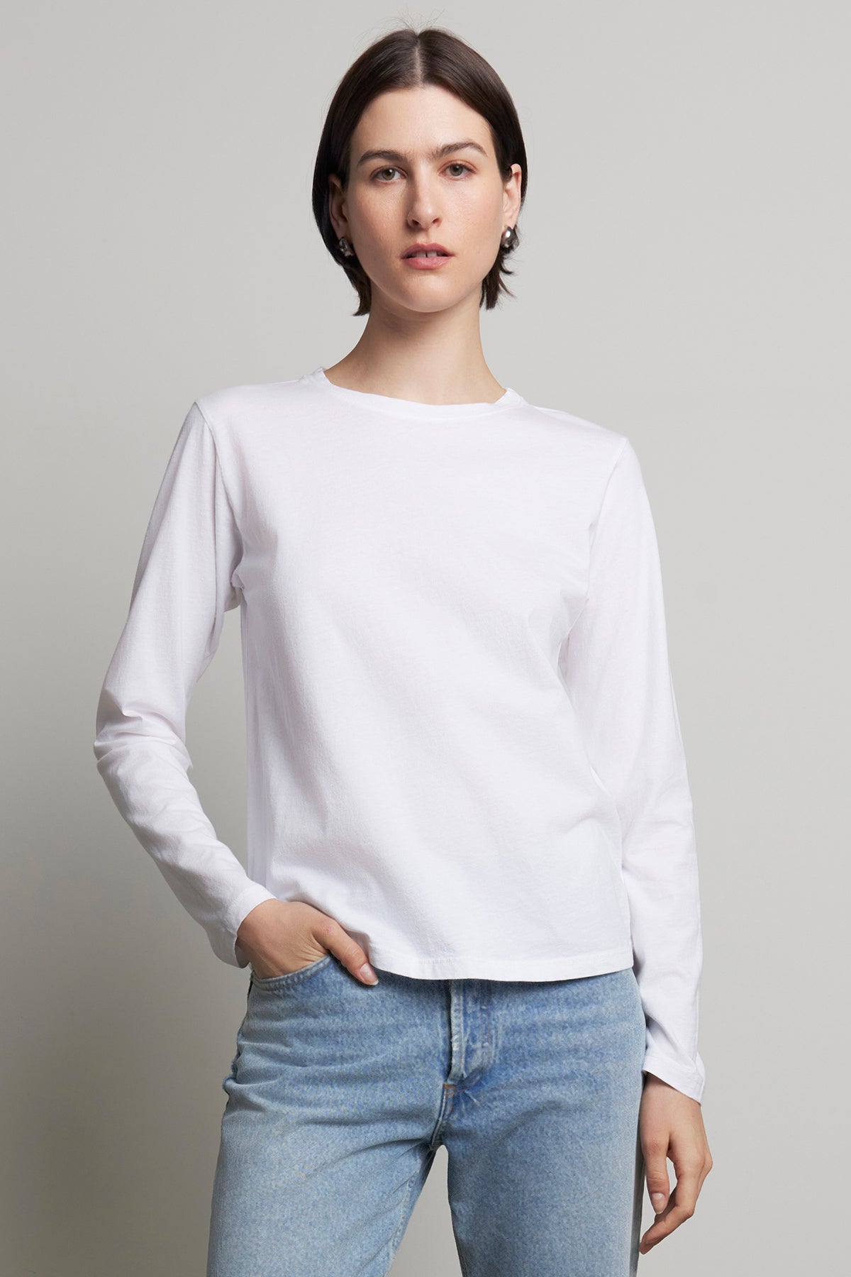 A person wearing the VICENTE TEE by Velvet by Jenny Graham, a relaxed-fit white long-sleeve shirt made from organic cotton, and blue jeans stands against a plain background.-38866409816257