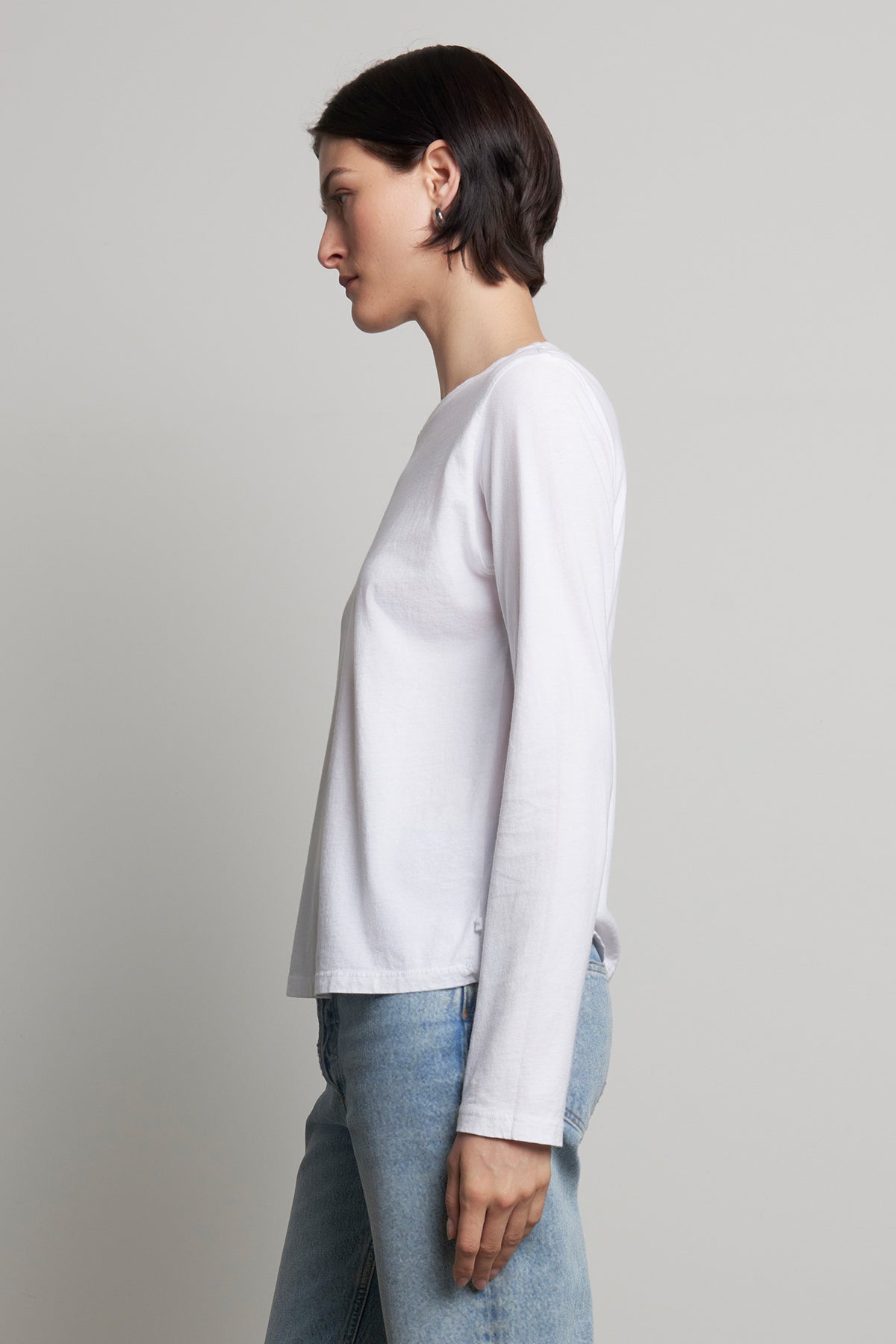   A person wearing the VICENTE TEE, a white long-sleeve organic cotton shirt from Velvet by Jenny Graham, and relaxed-fit blue jeans stands sideways. 