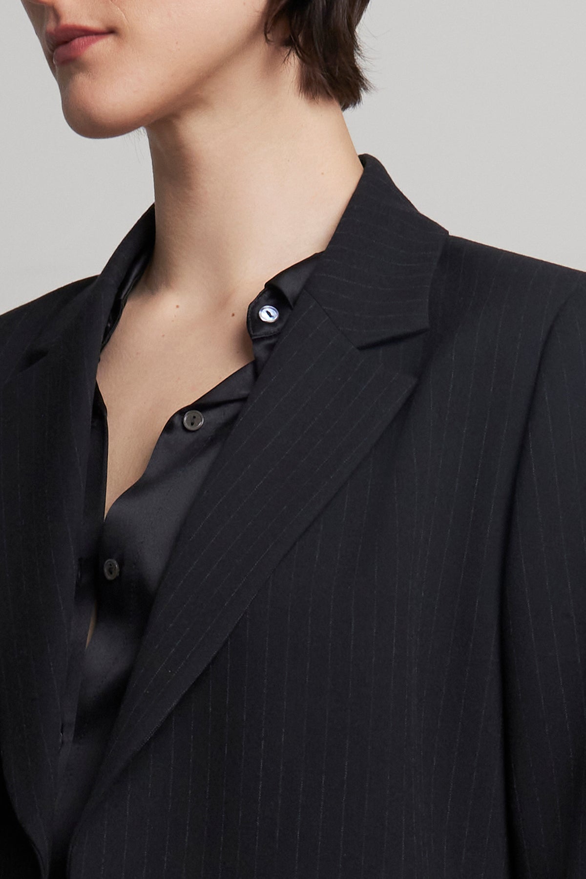   The individual is depicted from the chest up, dressed in a pinstripe polyester ensemble featuring a partially unbuttoned black shirt and the oversized FRESNO PINSTRIPE BLAZER by Velvet by Jenny Graham. The notched collar adds a sharp touch to the look. 