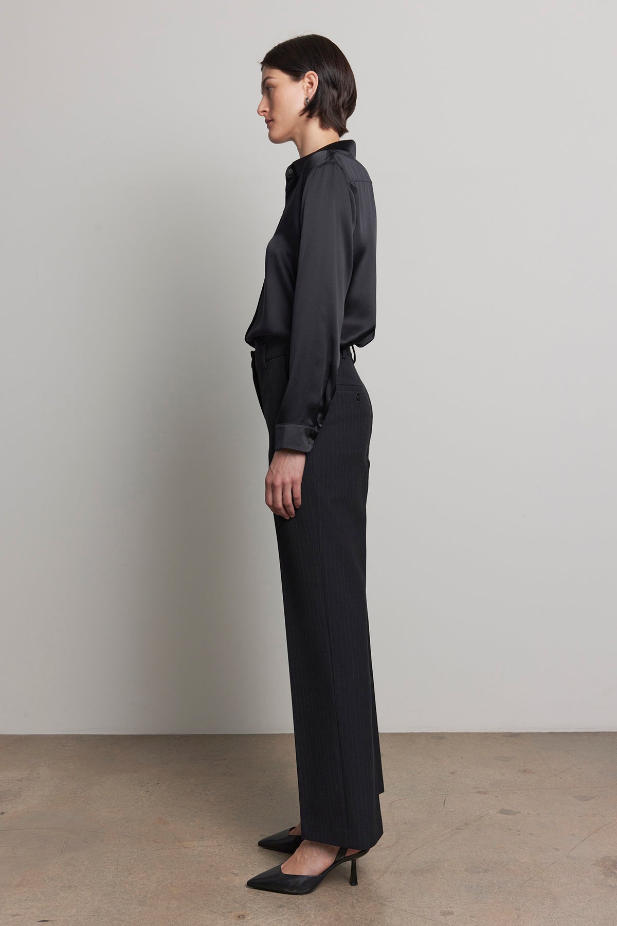   A person standing in profile, wearing a dark long-sleeved shirt paired with the TAHOE PINSTRIPE PANT by Velvet by Jenny Graham and complemented by black heels, against a neutral background. 