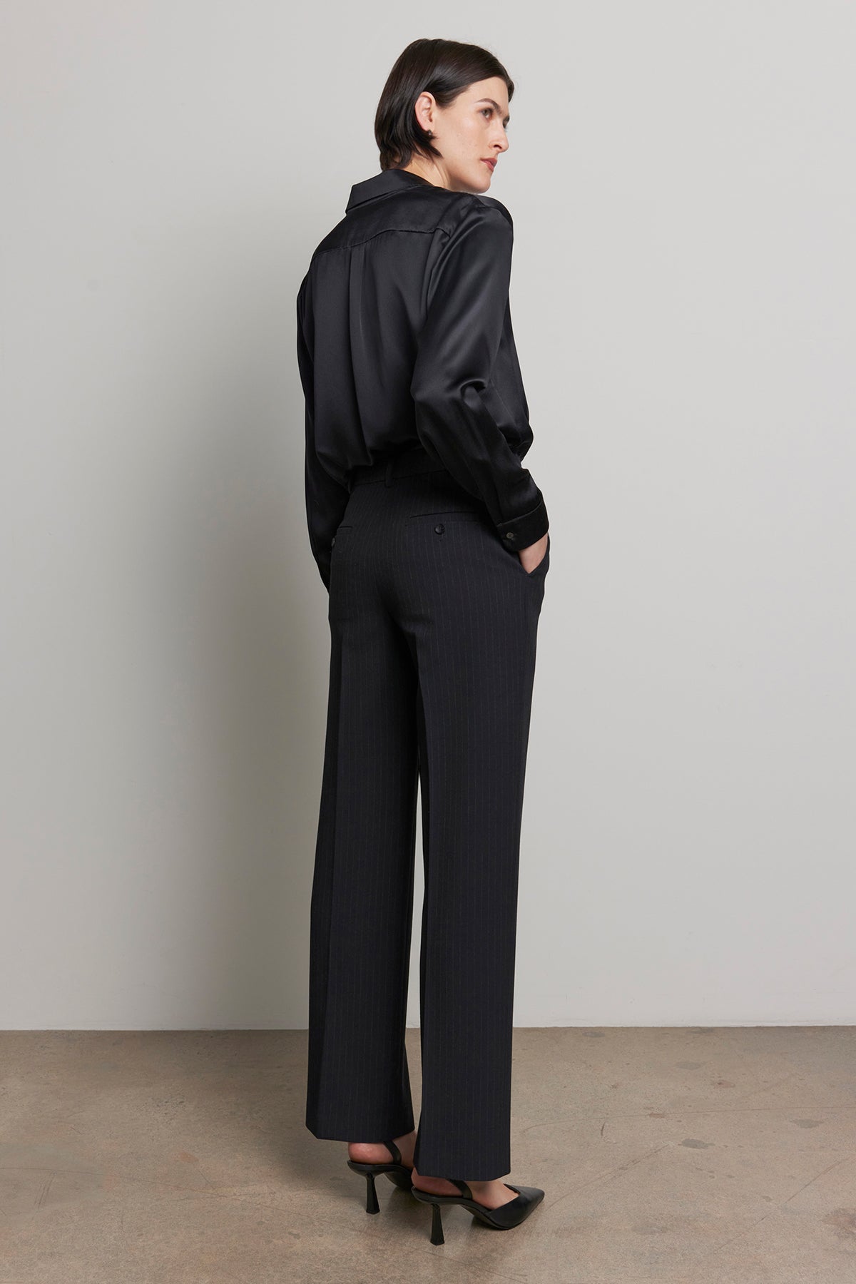   A person wearing a black silk shirt and the TAHOE PINSTRIPE PANT by Velvet by Jenny Graham stands sideways with hands in pockets against a plain gray background. 