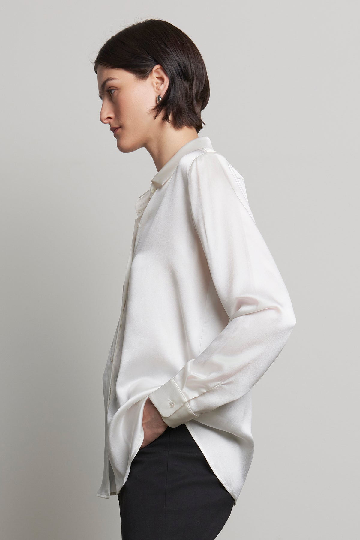   A person wearing the CALABASAS SILK CHARMEUSE BLOUSE by Velvet by Jenny Graham and black pants, with hands in pockets, is facing left against a gray background. 