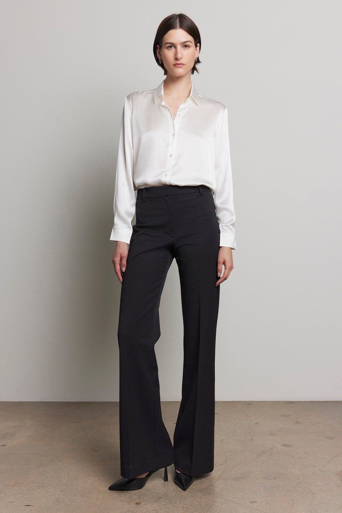   Against a plain background, a person is standing in the MARIPOSA PANT by Velvet by Jenny Graham, featuring a white blouse and black, soft suiting fabric with flared legs. 