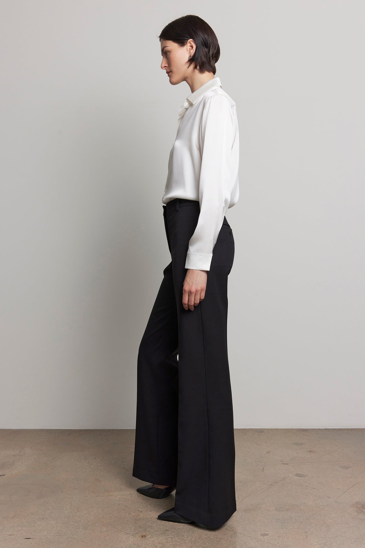   A person stands in profile wearing a white shirt and the MARIPOSA PANT by Velvet by Jenny Graham, featuring black soft suiting fabric with flared legs, against a plain background. 