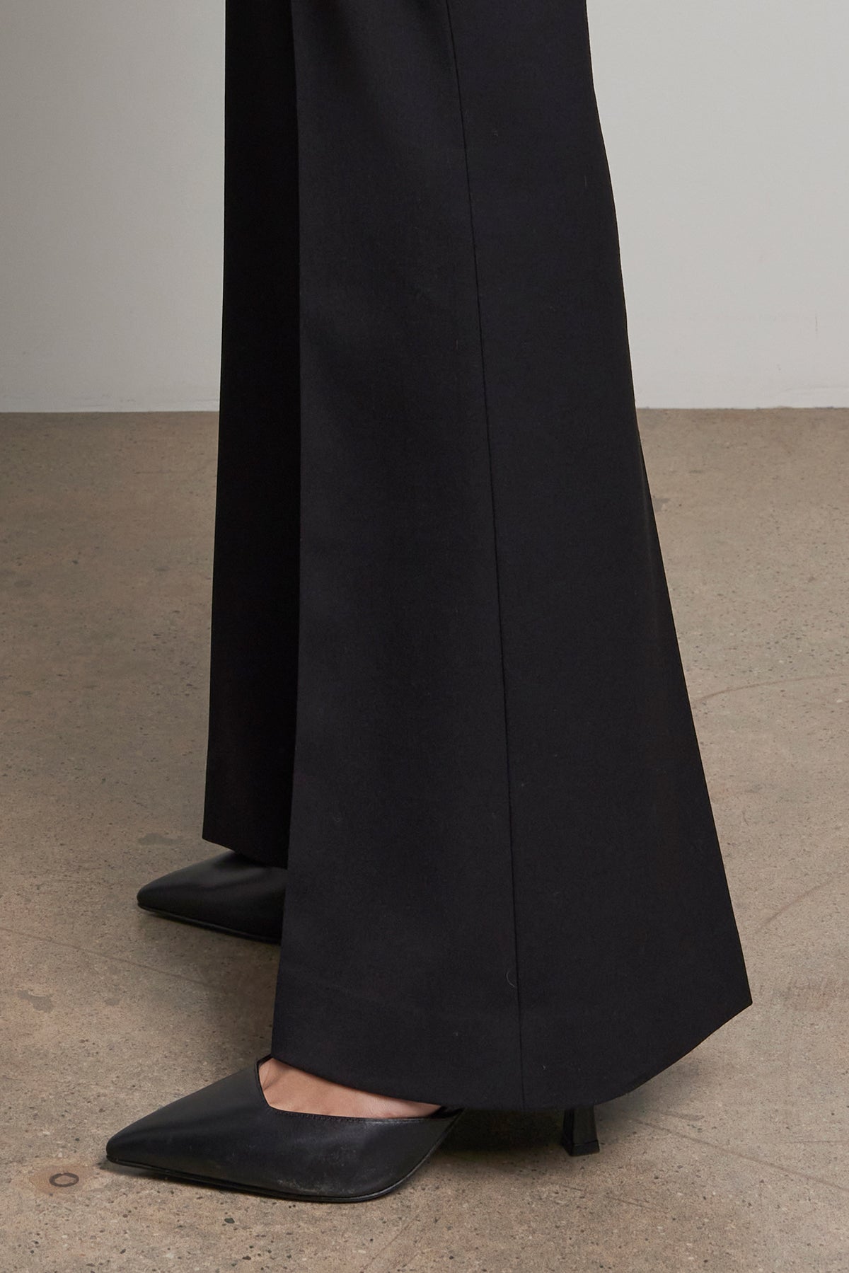   A person stands on a concrete floor wearing the MARIPOSA PANT by Velvet by Jenny Graham, which features black wide-leg pants with a tailored closure, paired with pointed black heels. 