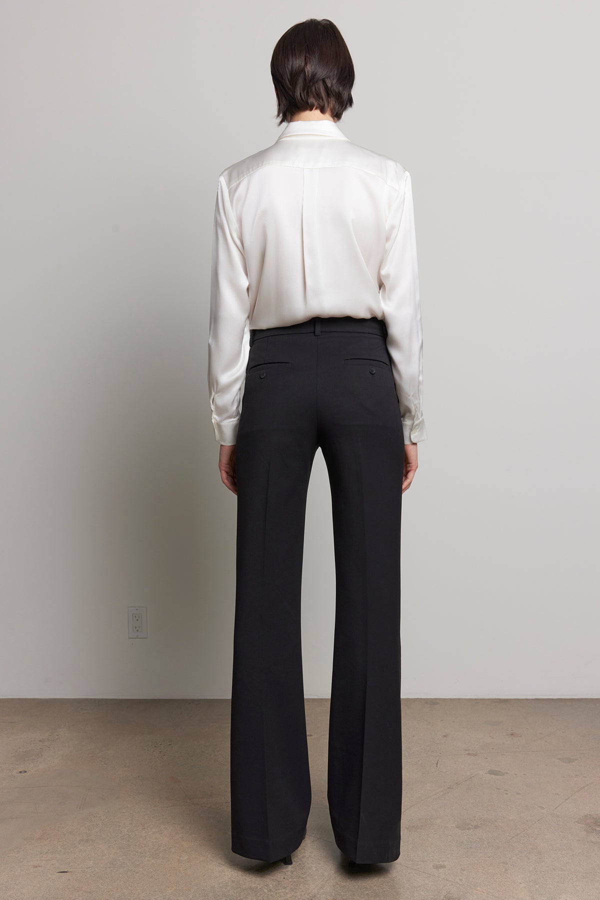   Back view of a person wearing soft suiting MARIPOSA PANT by Velvet by Jenny Graham and a white blouse, standing on a plain surface and facing a light gray wall. 