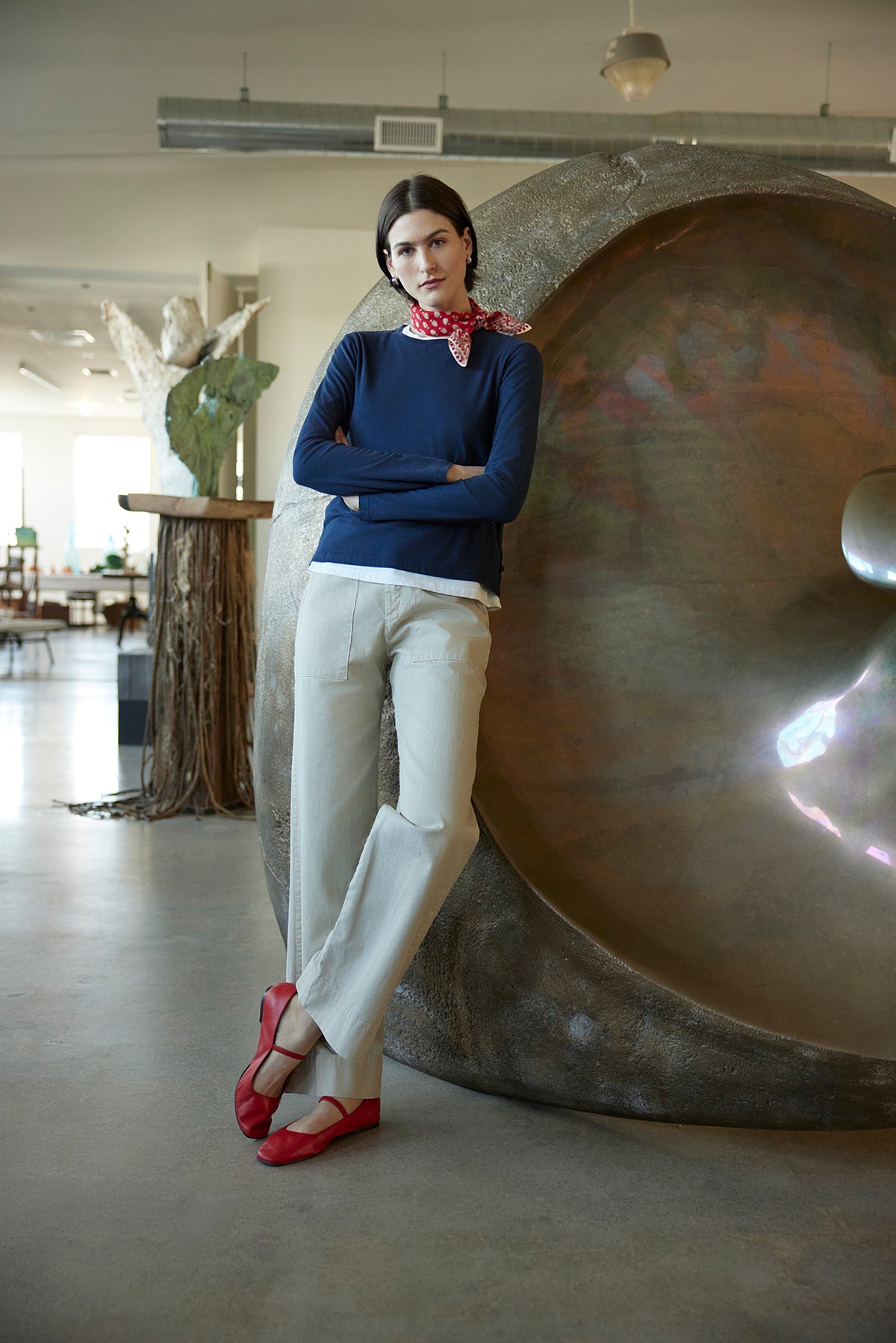   Dressed in a VICENTE TEE by Velvet by Jenny Graham, relaxed fit and navy long sleeve, with white pants and red shoes, a person leans against a large metallic sculpture indoors. 