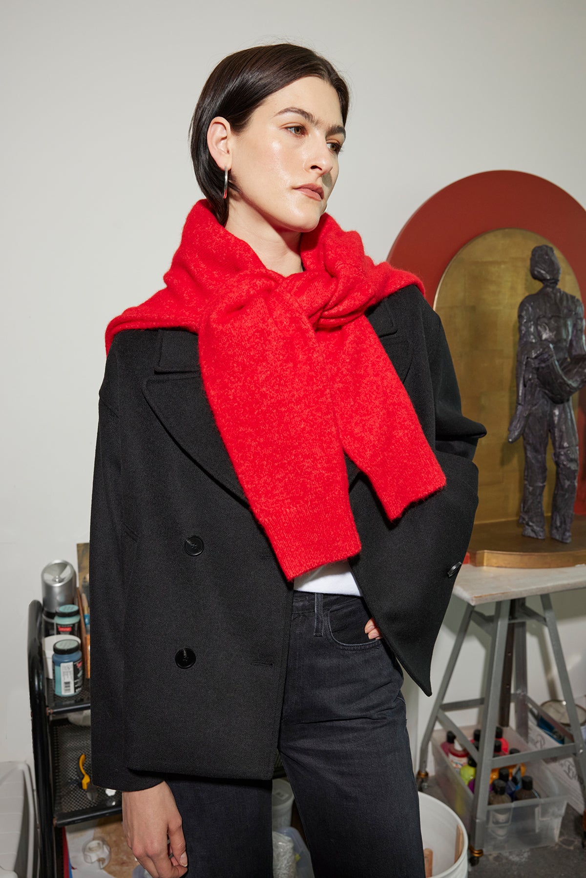 A person in a black jacket and red scarf stands in a room with art supplies and a sculpture in the background, their eyes scanning the scene. Nearby, the CYPRESS SWEATER by Velvet by Jenny Graham rests on an easel, its warm tones complementing the creative energy of the space.-38318581383361