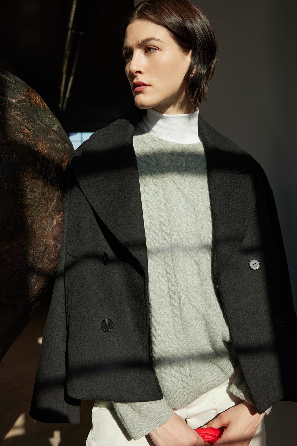   A person wearing the WILSHIRE SWEATER by Velvet by Jenny Graham and a black coat stands in the sunlight, looking away. 