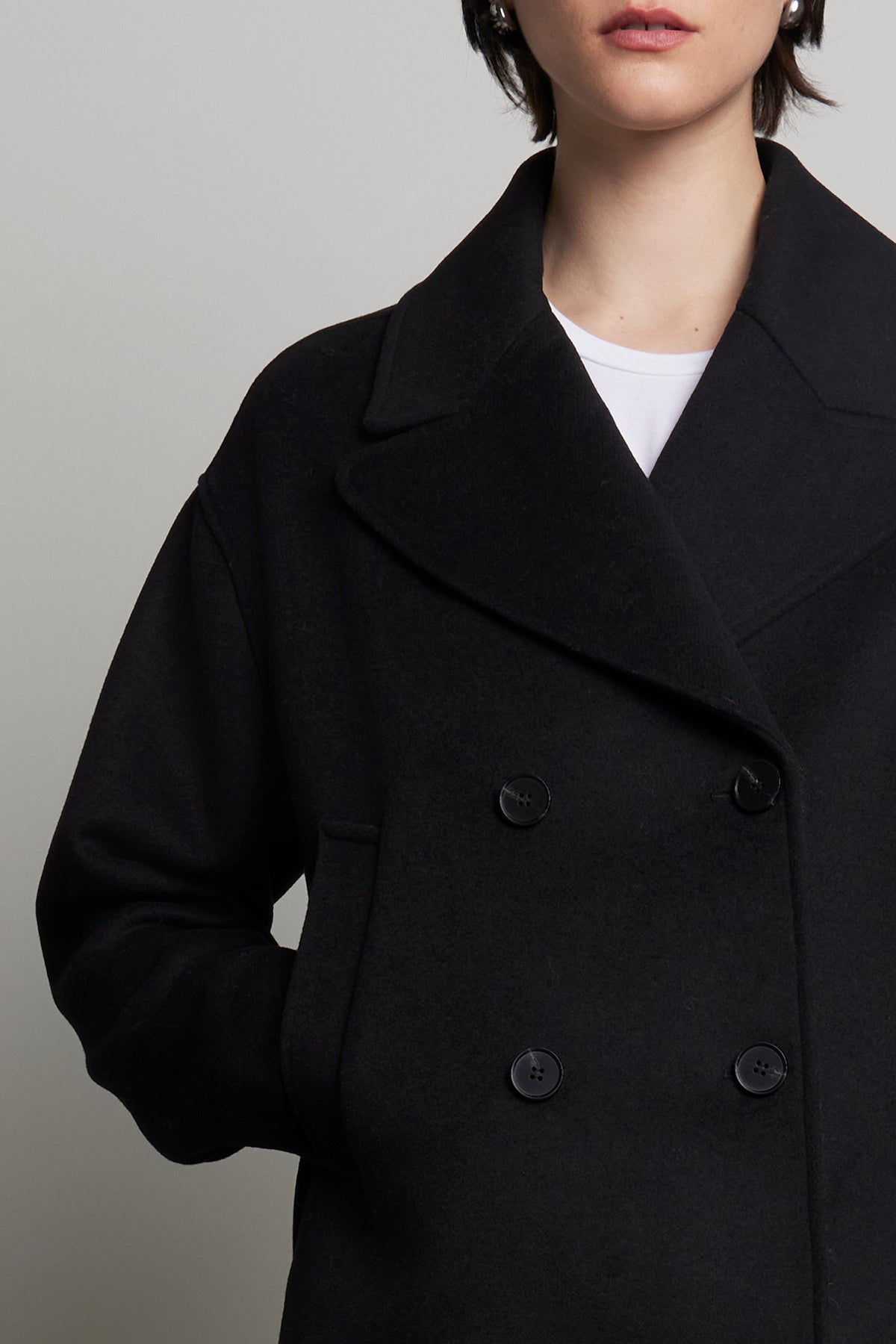   Against a plain backdrop, an individual stands with hands in pockets, donning the WESTWOOD PEACOAT from Velvet by Jenny Graham. This black double-breasted peacoat, made from a wool blend, radiates timeless appeal. 