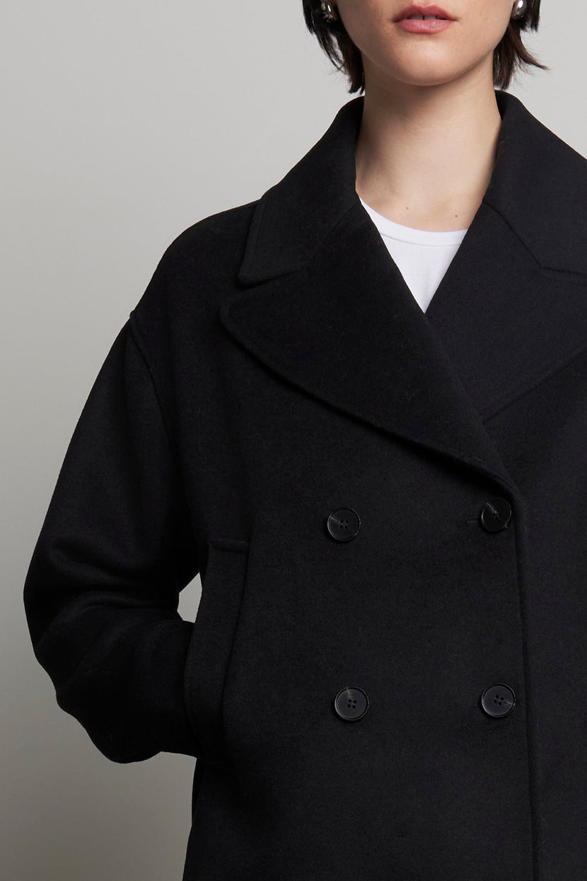 Against a plain backdrop, an individual stands with hands in pockets, donning the WESTWOOD PEACOAT from Velvet by Jenny Graham. This black double-breasted peacoat, made from a wool blend, radiates timeless appeal.