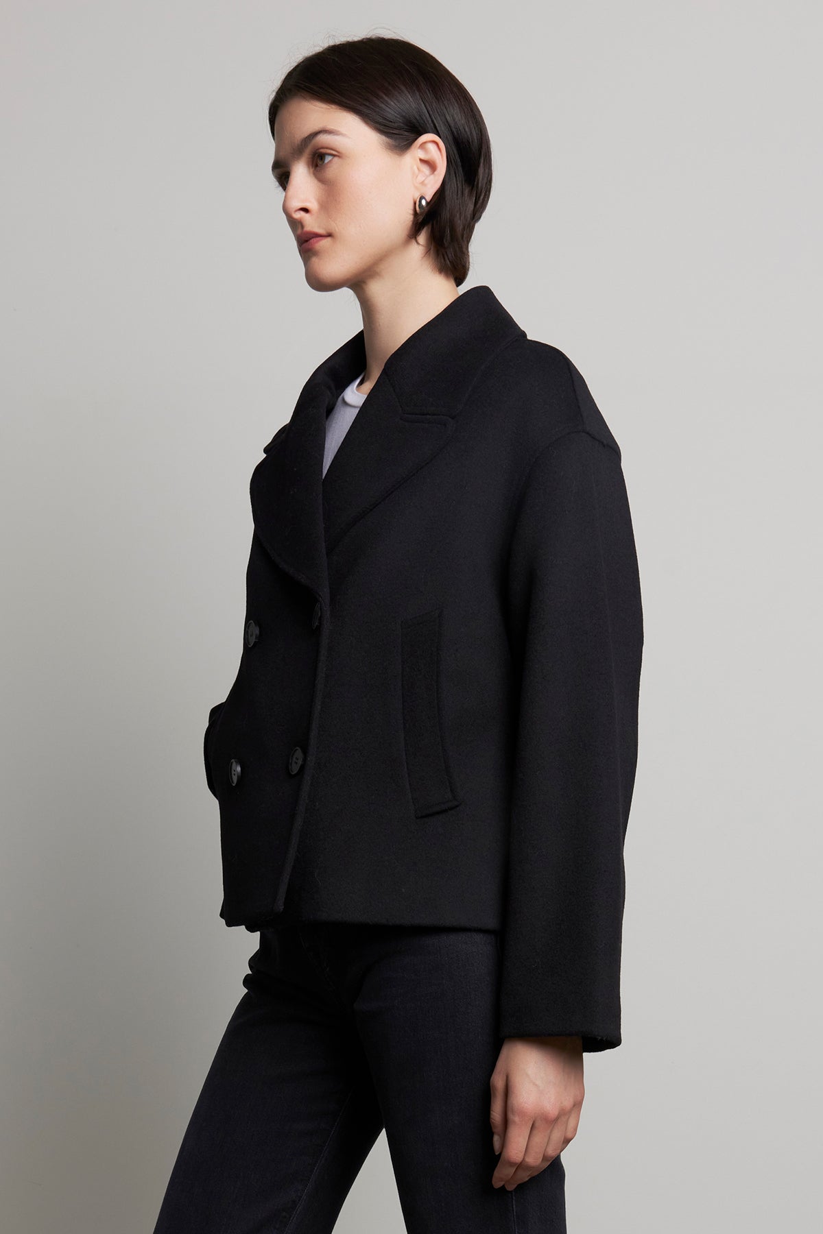 Dressed in the WESTWOOD PEACOAT by Velvet by Jenny Graham, paired with black jeans, a person stands sideways against a plain background, exuding timeless appeal.-38318587183297