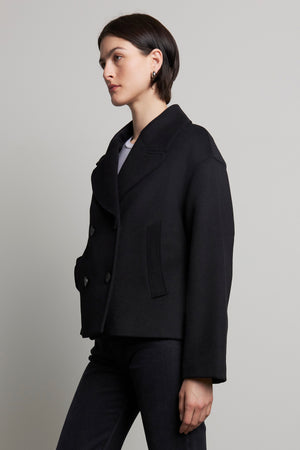 Dressed in the WESTWOOD PEACOAT by Velvet by Jenny Graham, paired with black jeans, a person stands sideways against a plain background, exuding timeless appeal.