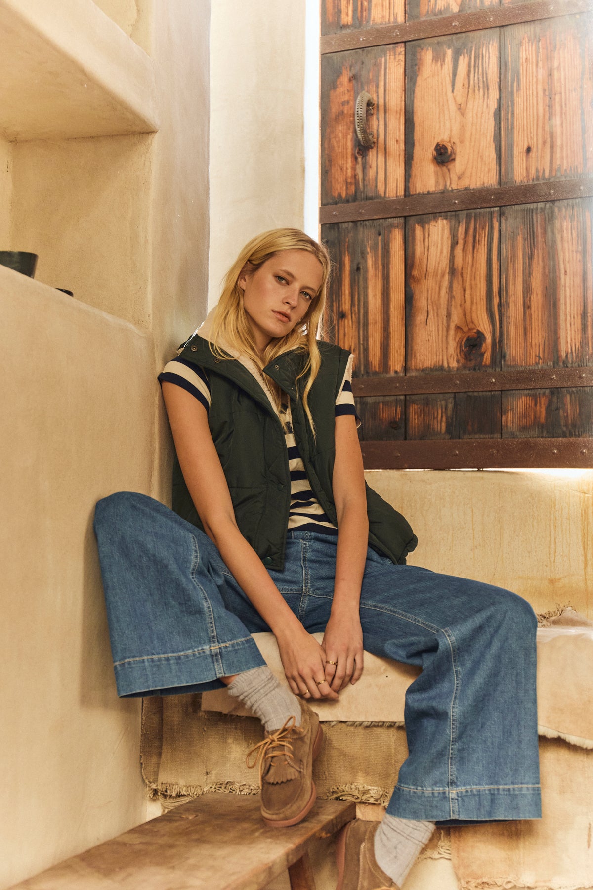 A woman sits on indoor stone steps wearing a striped shirt, wide-leg jeans, socks, brown shoes, and the CATHY PUFFER SHERPA REVERSIBLE VEST by Velvet by Graham & Spencer. A wooden door is in the background.-39744837222593