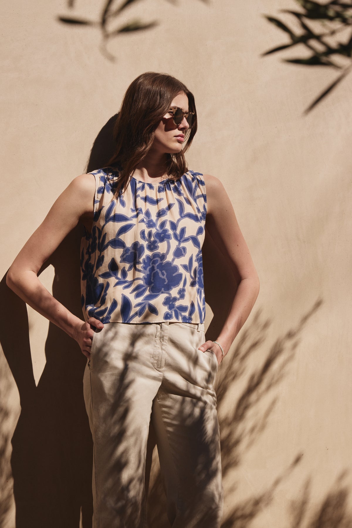 Wearing Velvet by Graham & Spencer's KATERINA TOP, a person in sunglasses stands against a beige wall with hands in pockets, surrounded by partial shadows and plant leaves.-38928816996545