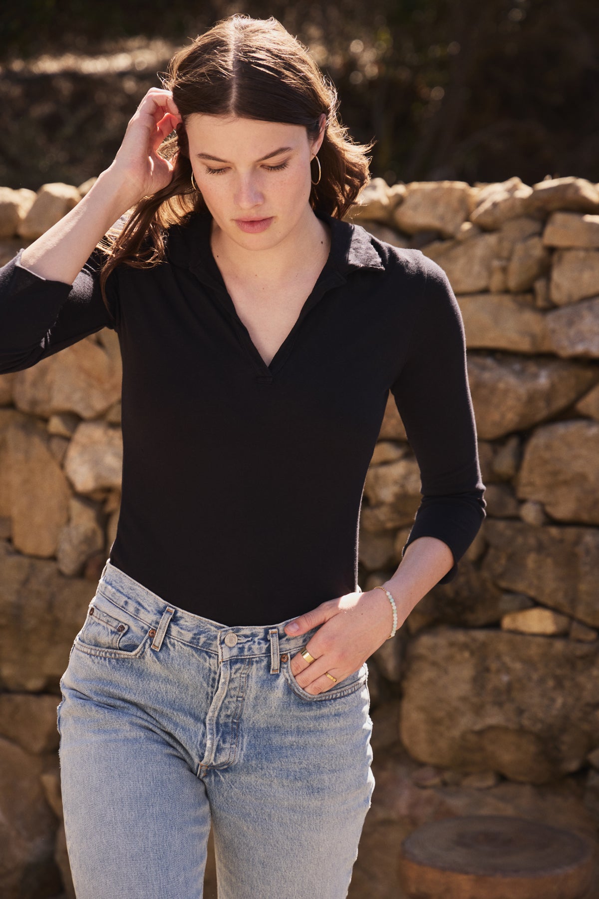 Wearing a REED POLO by Velvet by Graham & Spencer with jeans, a person touches their hair outdoors against a stone wall backdrop.-38928823484609
