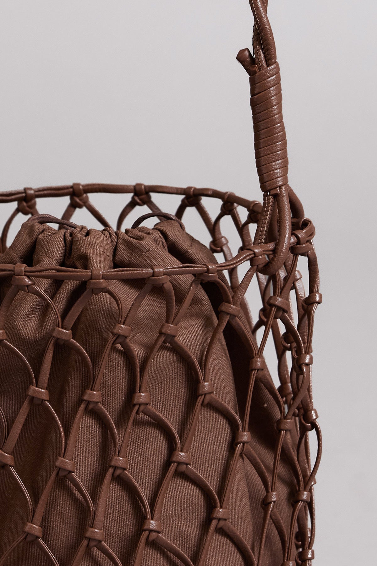   Close-up of a brown Velvet by Graham & Spencer mesh tote bag with a vegan leather wrapped handle against a neutral background. 