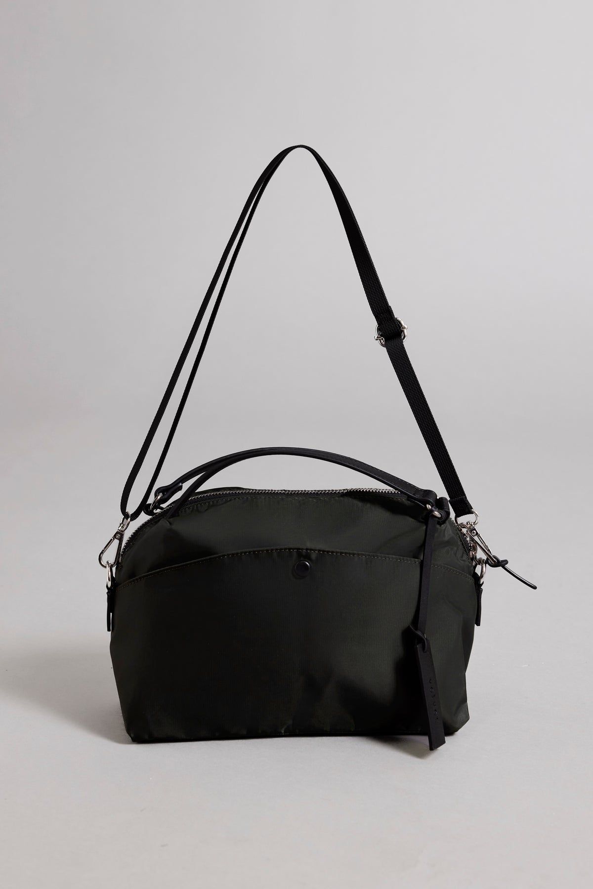   The NYLON CROSSBODY BAG by Velvet by Graham & Spencer is a stylish black handbag made from durable nylon, featuring an adjustable shoulder strap and a top handle. The design is finished with a zipper closure, all set against a simple gray background. 