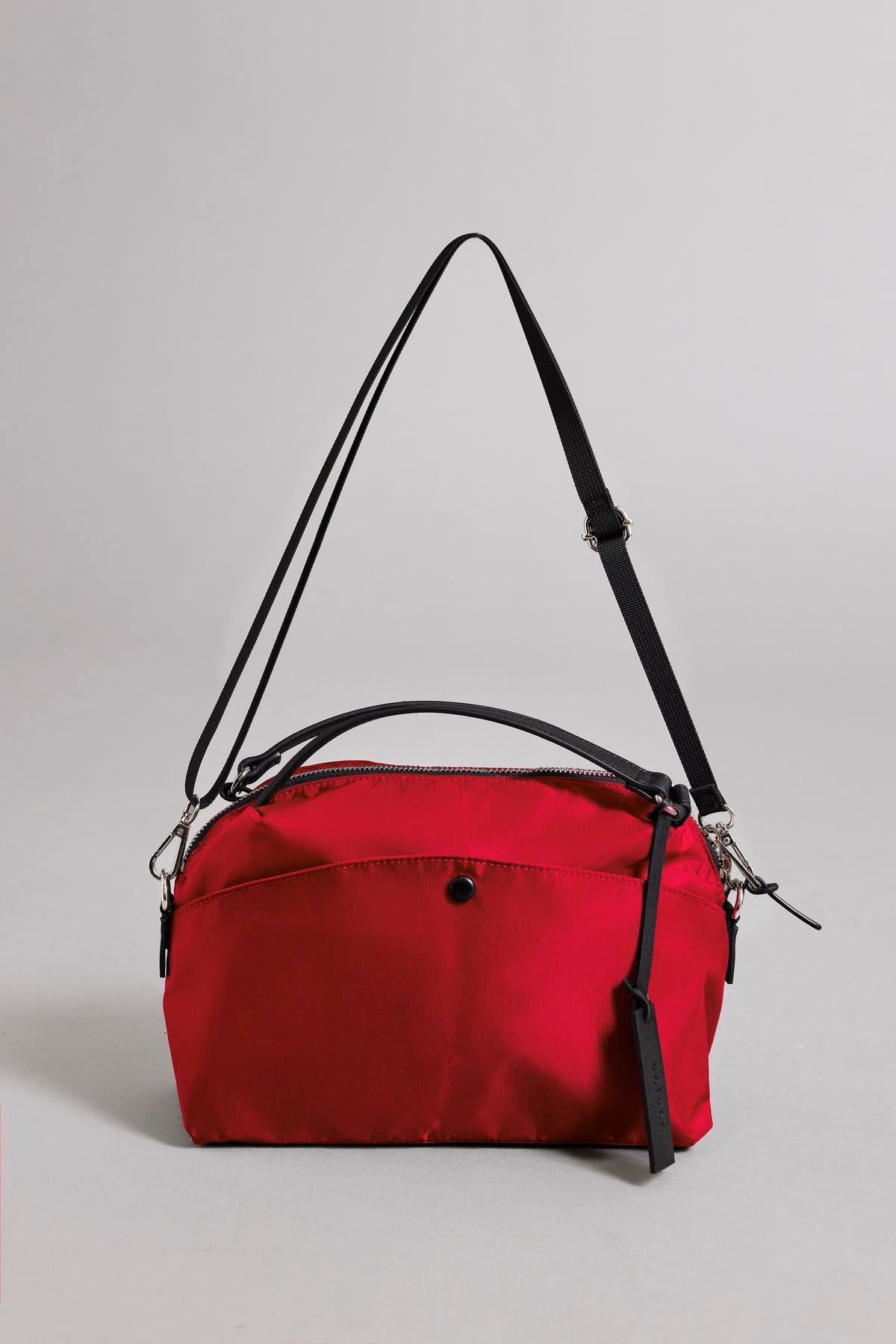   NYLON CROSSBODY BAG by Velvet by Graham & Spencer is a red handbag made from durable nylon, featuring a black strap, zipper closure, and a small black tag. It is perfect for everyday use and is showcased on a plain gray background. 