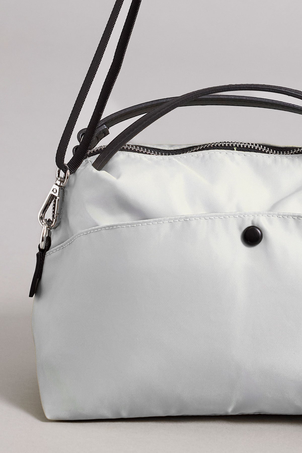   A close-up of a light gray NYLON CROSSBODY BAG by Velvet by Graham & Spencer, made from durable nylon, featuring a secure zipper closure, a black button on the front, and black straps. 