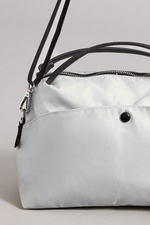 A close-up of a light gray NYLON CROSSBODY BAG by Velvet by Graham & Spencer, made from durable nylon, featuring a secure zipper closure, a black button on the front, and black straps.