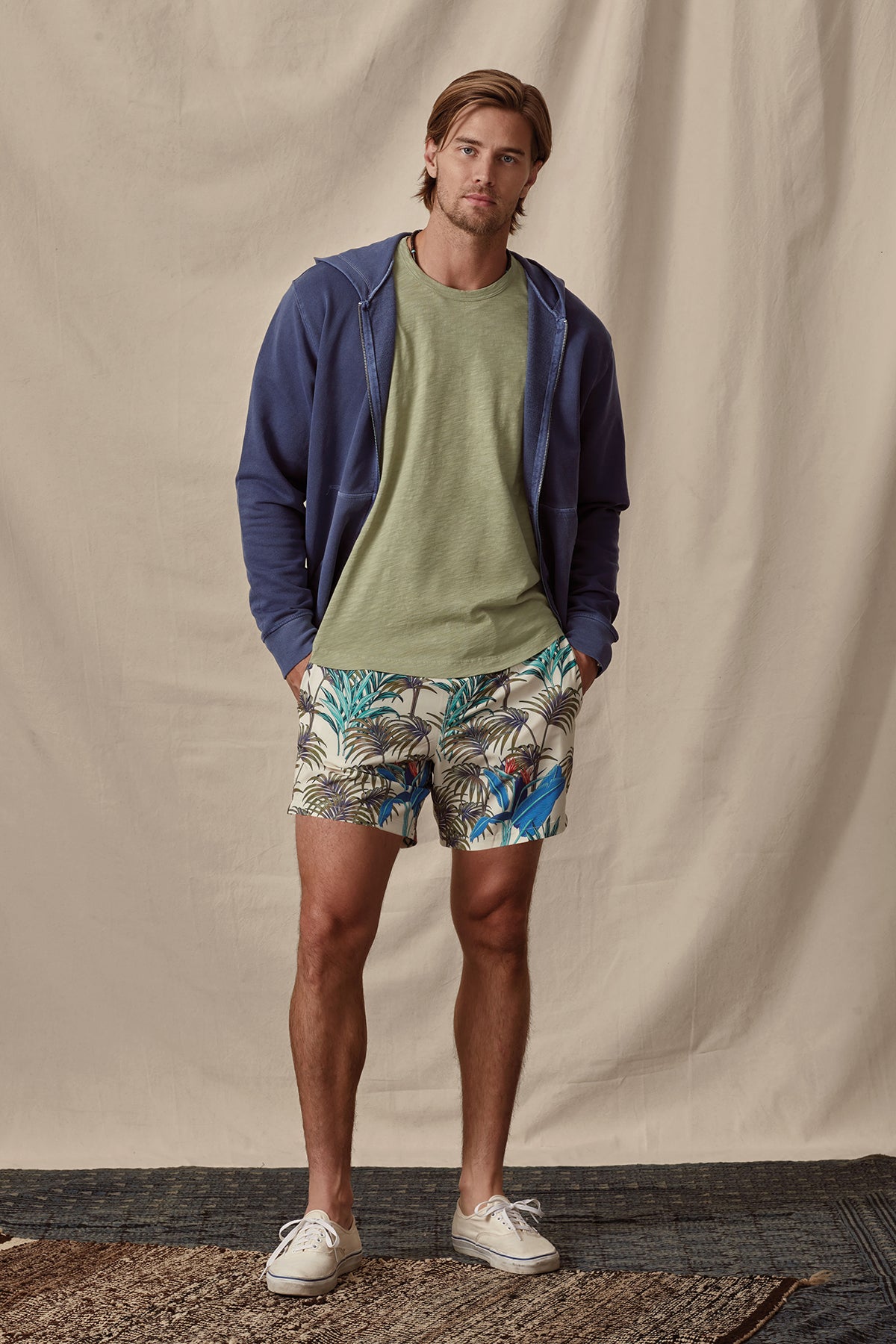   A man wearing a zip-up hoodie, t-shirt, and RICARDO SWIM SHORT by Velvet by Graham & Spencer stands confidently against a cream backdrop. 