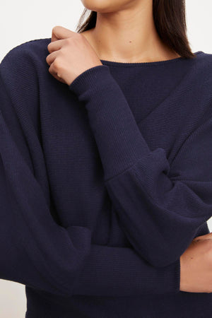The model is wearing a Velvet by Graham & Spencer navy sweater with a ruffled sleeve called the MACKAY BRUSHED RIB TOP.