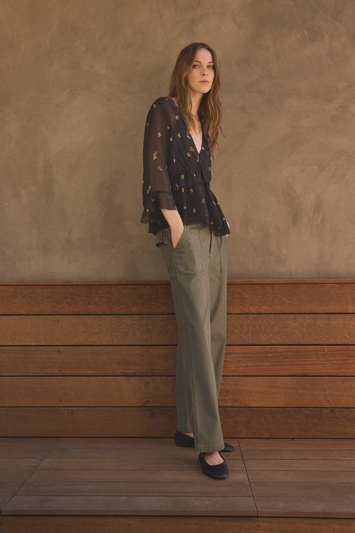 A woman stands against a brown textured wall with her hands in her pockets, wearing a sheer black star-patterned blouse, the JACLYN PANT by Velvet by Graham & Spencer in olive green cotton sateen wide-leg style, and black shoes.